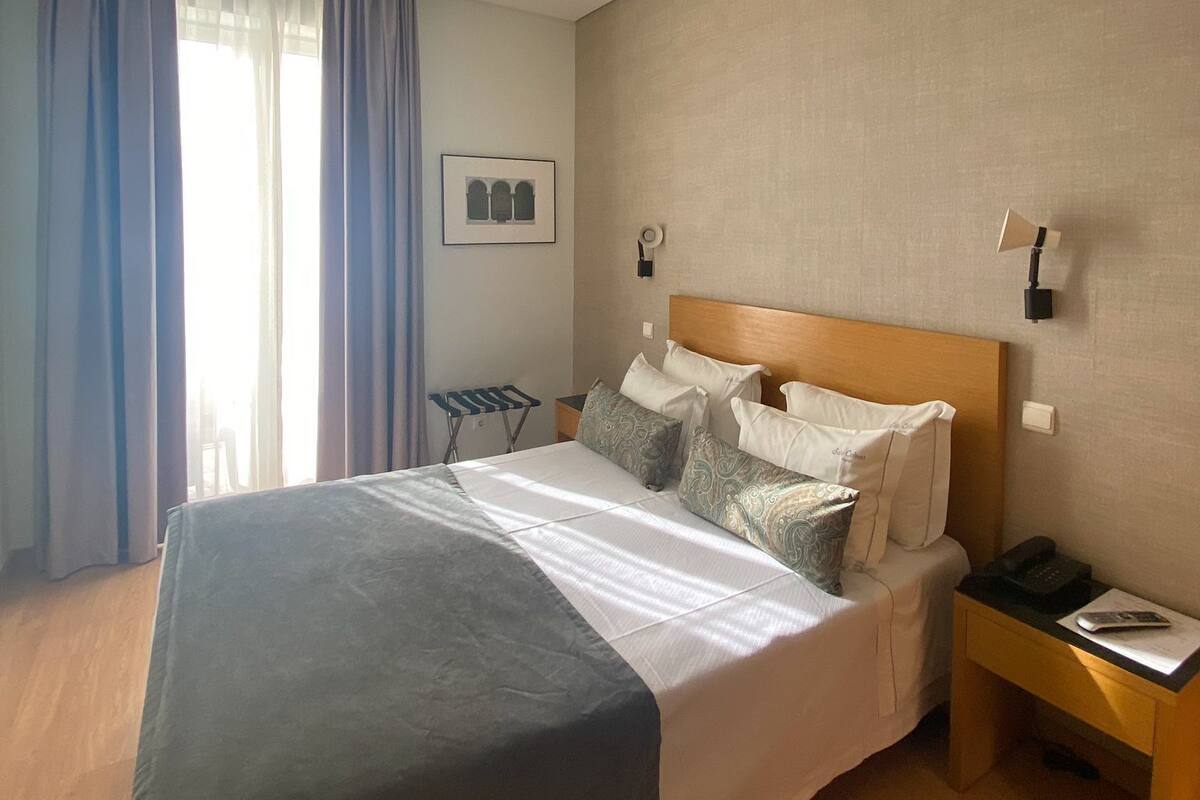 A neatly made bed with a grey blanket, a wooden headboard, and multiple pillows. There is a bedside table with a phone and a lamp. The room has a large window with blue curtains, a small table, and a wall-mounted picture.