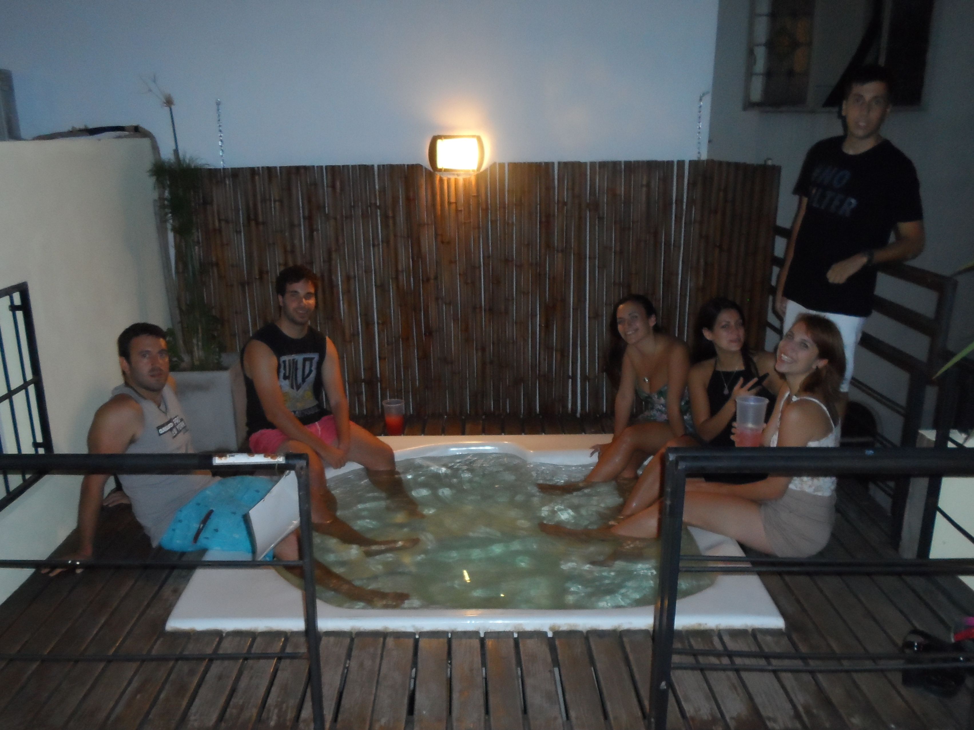 A group of people sitting around a hot tub in an outdoor setting with a wooden fence and a wall-mounted light.