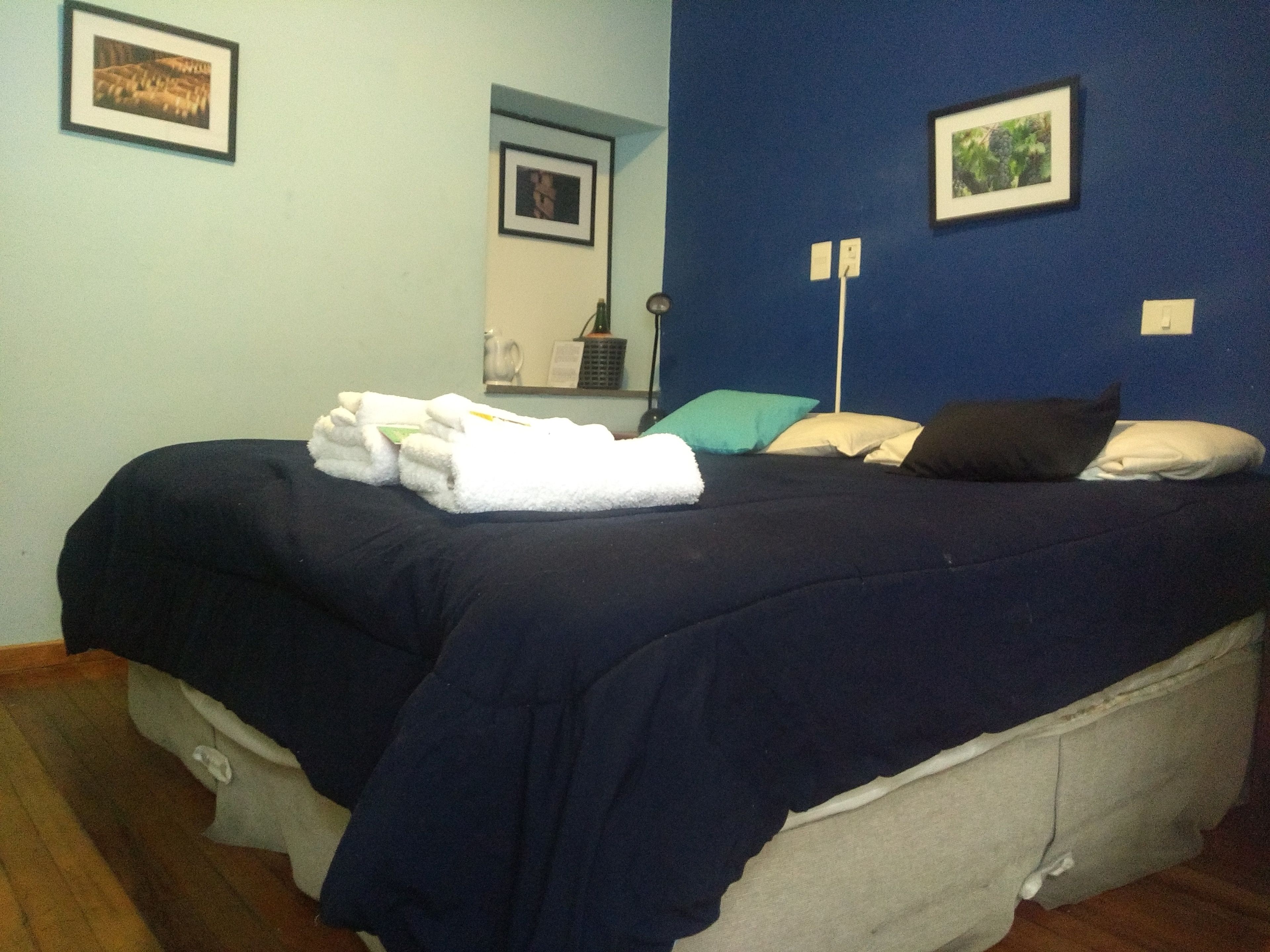 A neatly made bed with a dark blue bedspread and white towels, a teal pillow, and a dark brown pillow on a wooden floor.