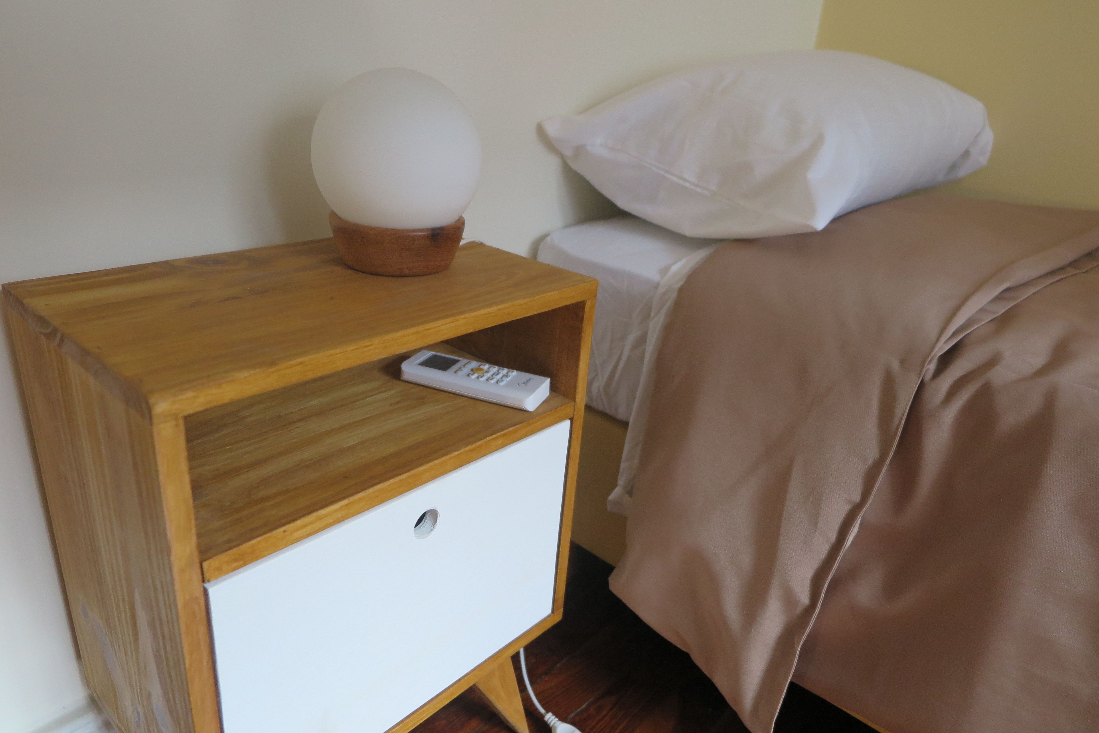 A wooden bedside table with a remote control, a white drawer, and a white pillow on a bed with a brown blanket.