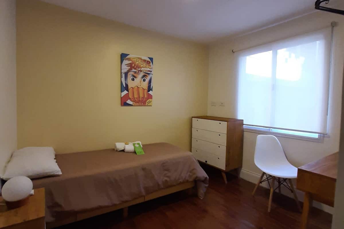 Ein kleiner, gut beleuchteter Raum mit einem Bett, einem Schreibtisch, einem Stuhl, einer Kommode und einem Bild an der Wand.
