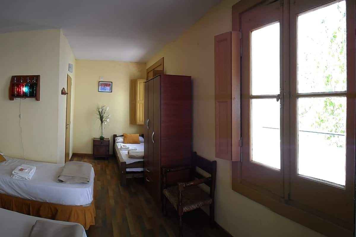 Ein Hotelzimmer mit zwei Betten, einem Holzschrank, einem kleinen Tisch und einem Fenster mit Blick auf Grünflächen.