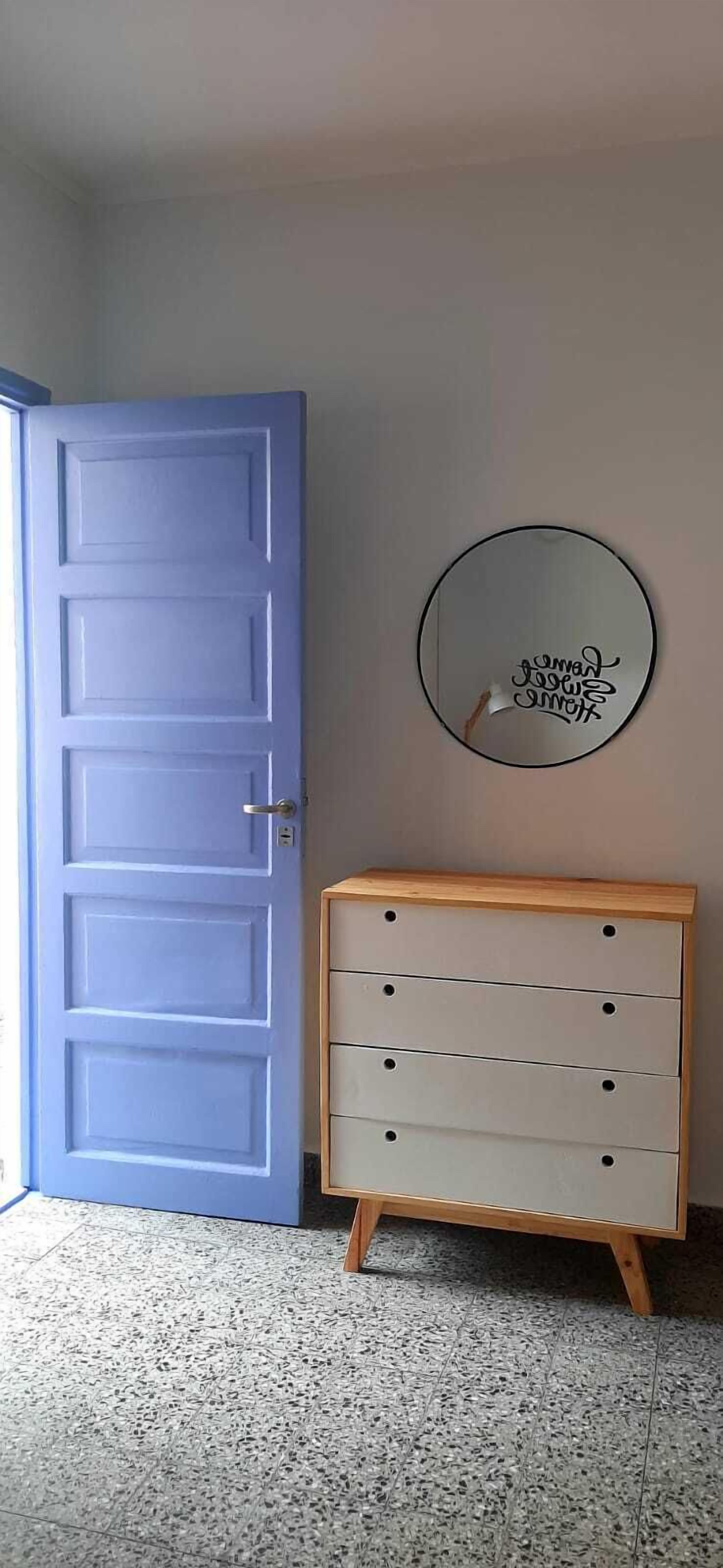 Una habitación con una puerta azul, un aparador de madera con cajones y un espejo redondo en la pared.