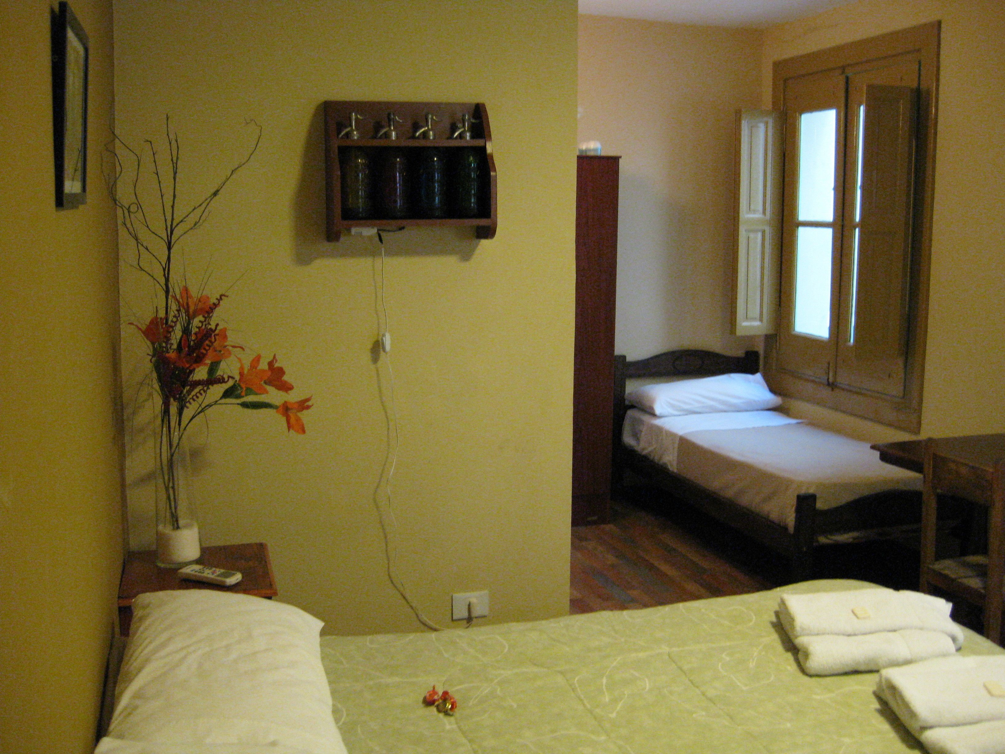 A hotel room with two beds, a small table with a vase of flowers, a mirror, and a shelf with bottles.