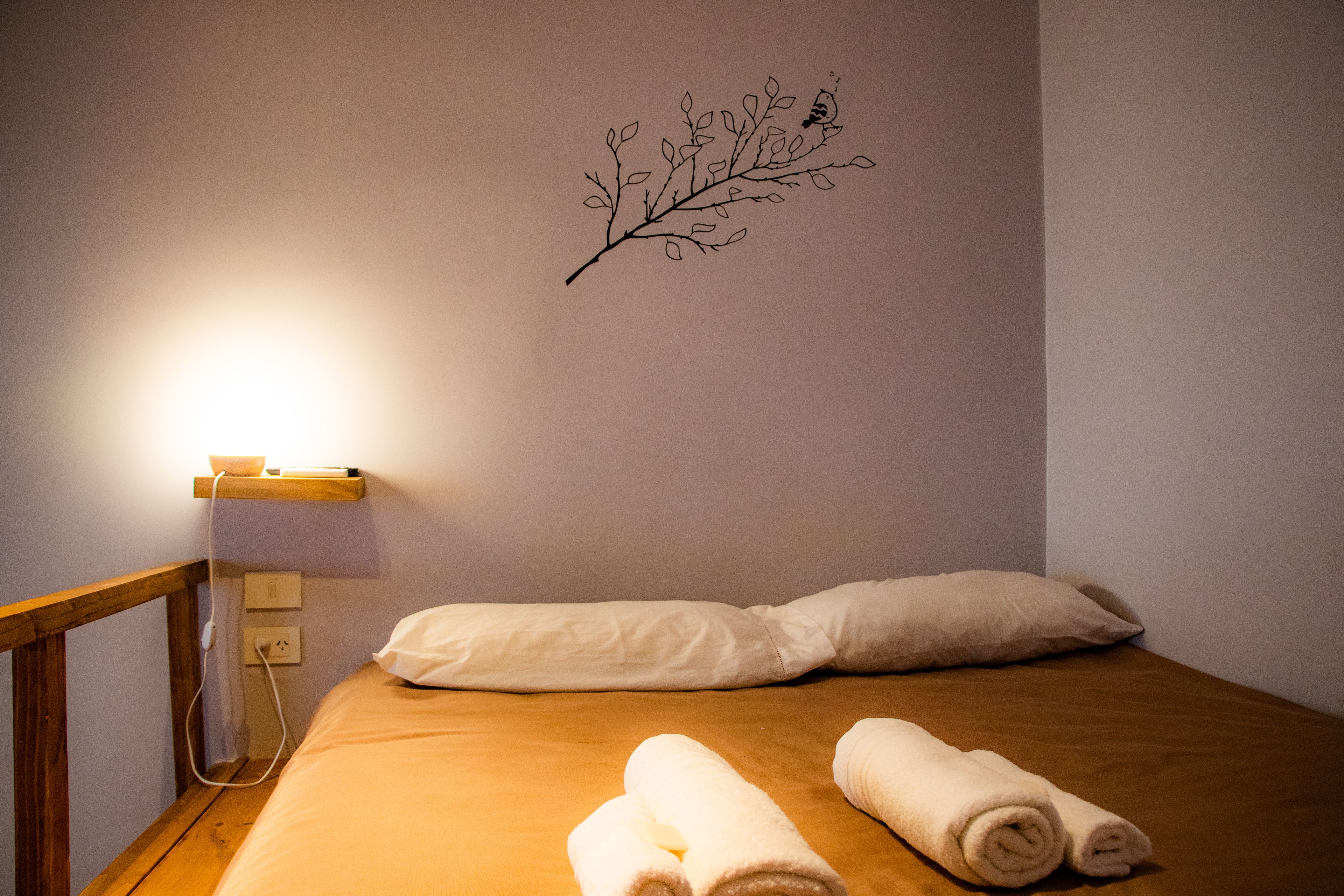 A neatly made bed with two pillows and two rolled towels, a wall-mounted branch decoration, and a bedside table with a lamp.