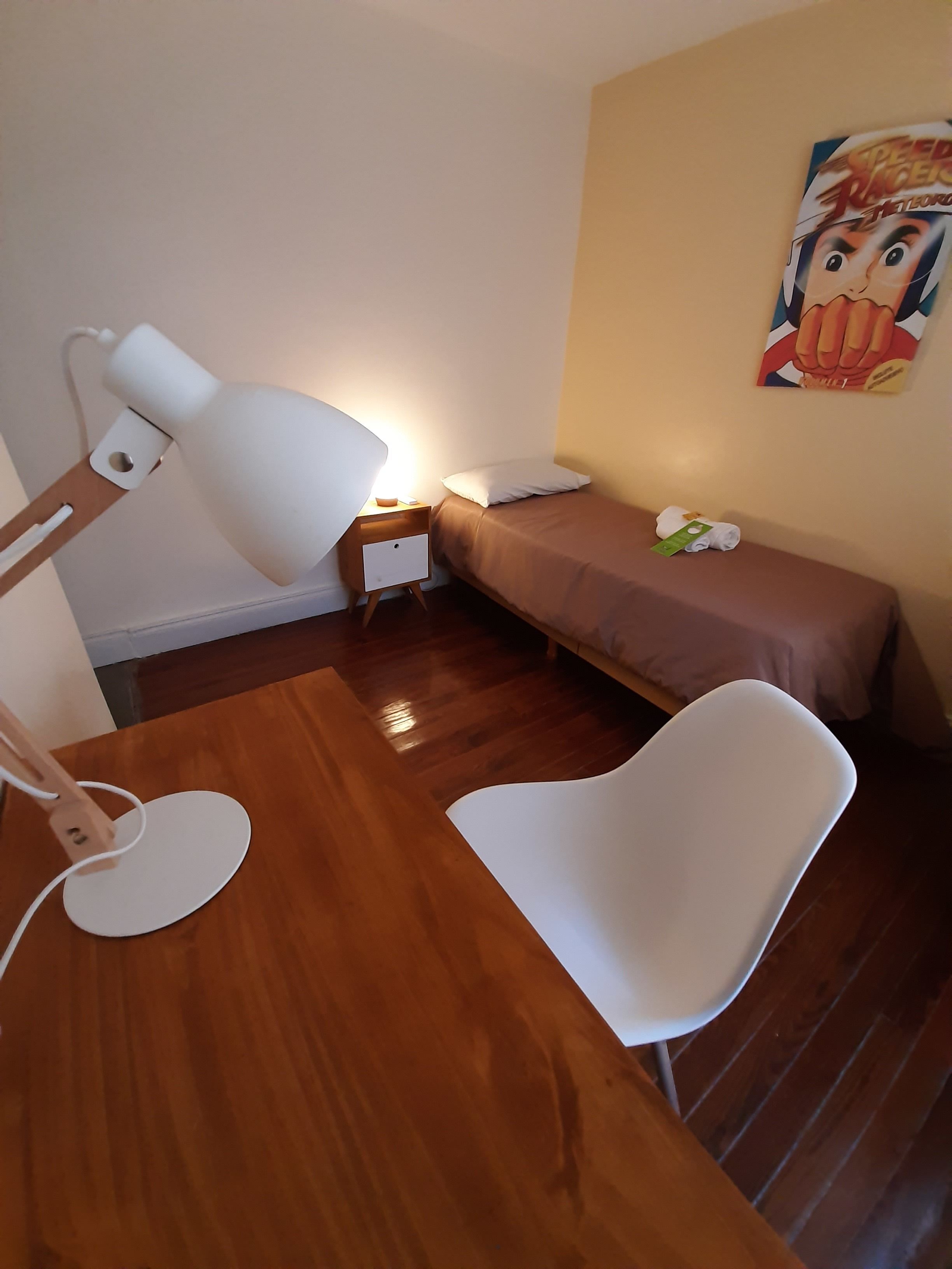 A small, neatly arranged bedroom with a wooden floor, a white desk with a lamp, a single bed with a brown mattress, and a wall-mounted poster.