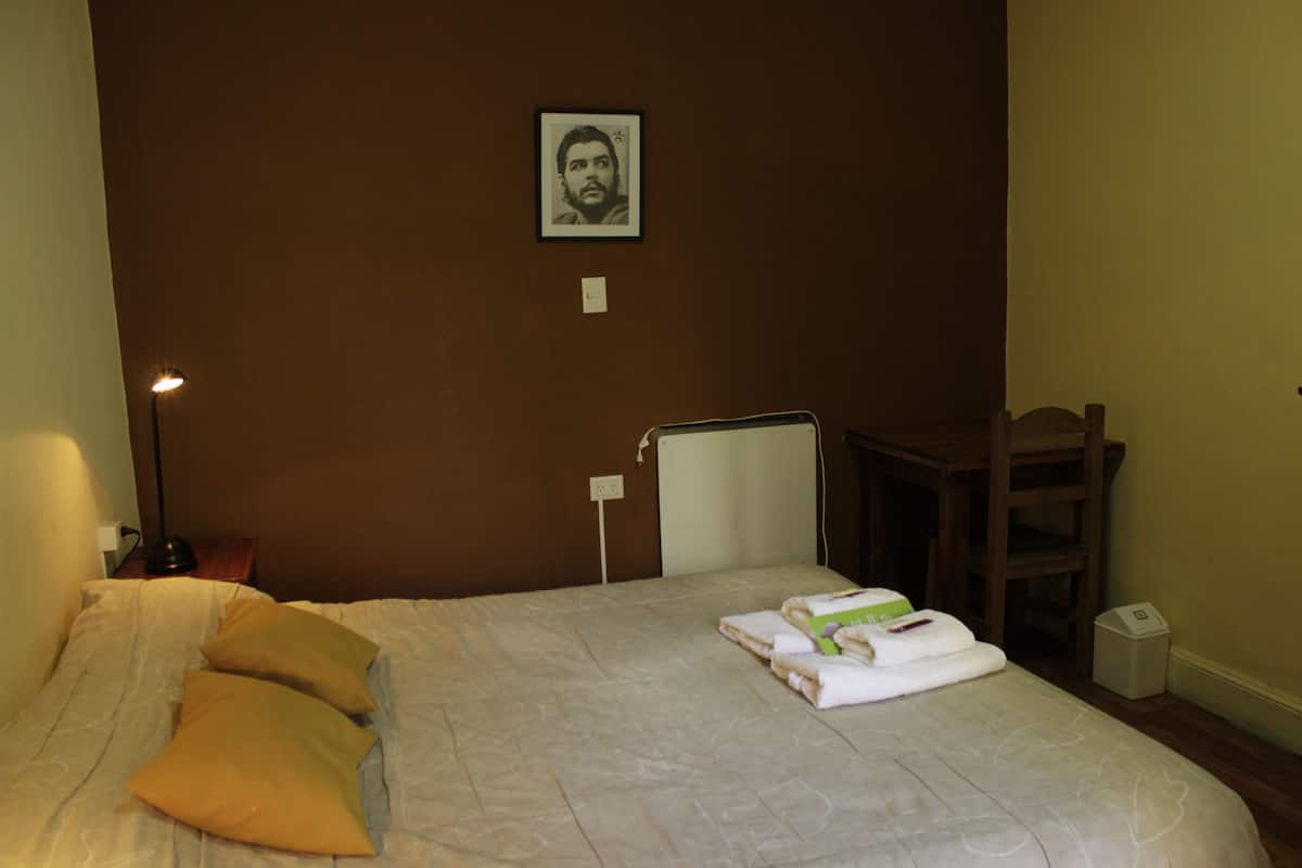 Ein ordentlich bezogenes Bett mit Kissen, eine Nachttischlampe, ein gerahmtes Bild an der Wand und ein kleiner Tisch mit einem Stuhl.