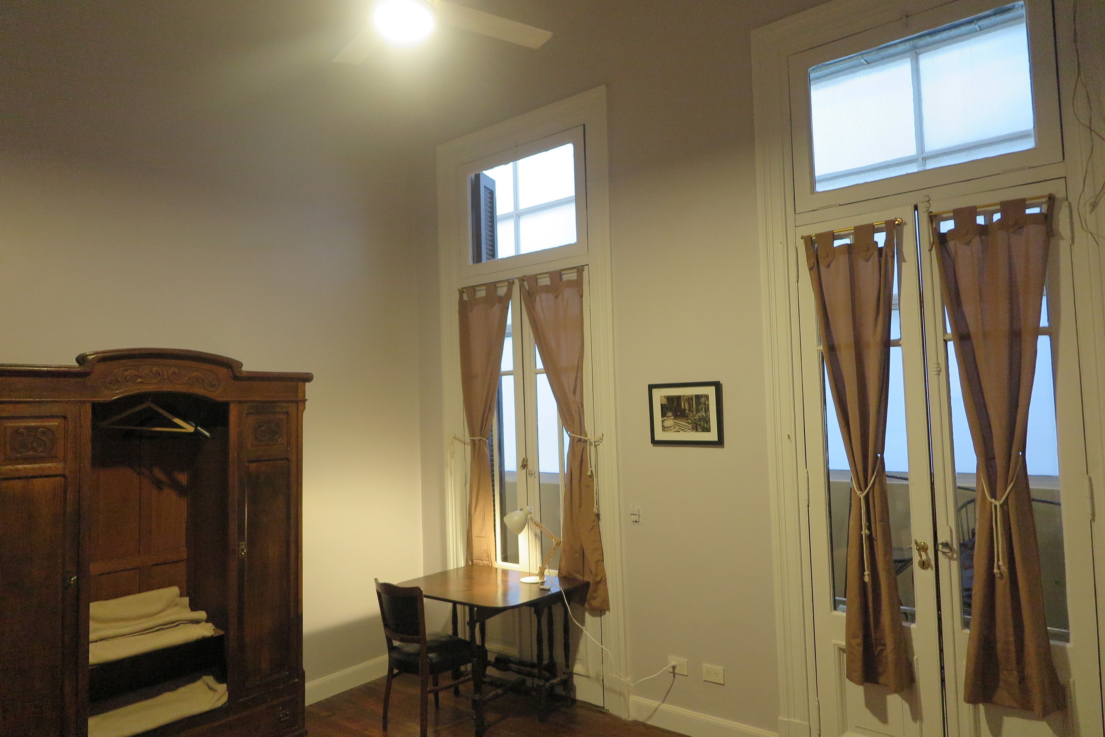 Una habitación con un armario de madera, una mesa con una lámpara y dos ventanas con cortinas.