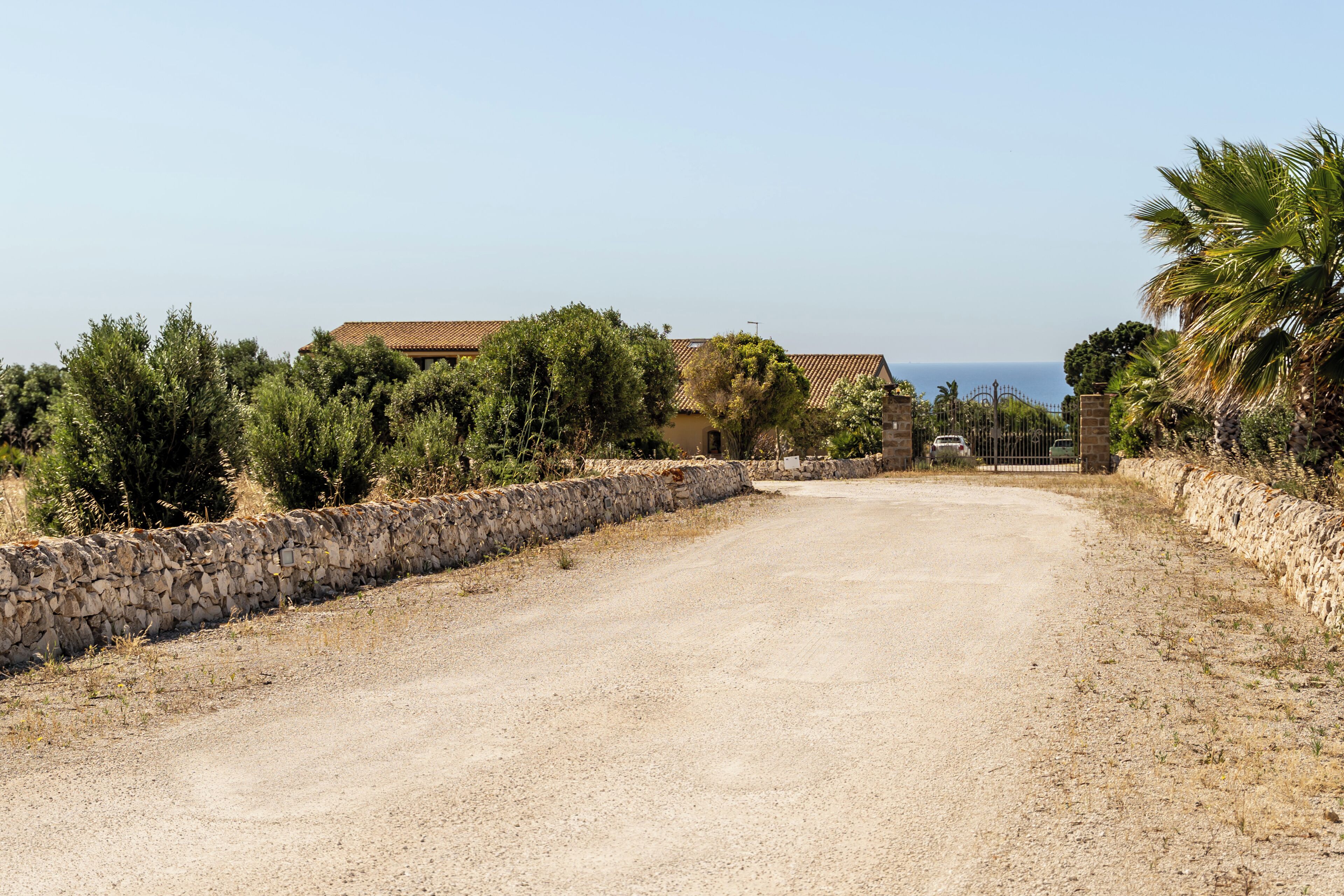 A gravel driveway leading to a stone wall with a gate, surrounded by vegetation and buildings with tiled roofs.