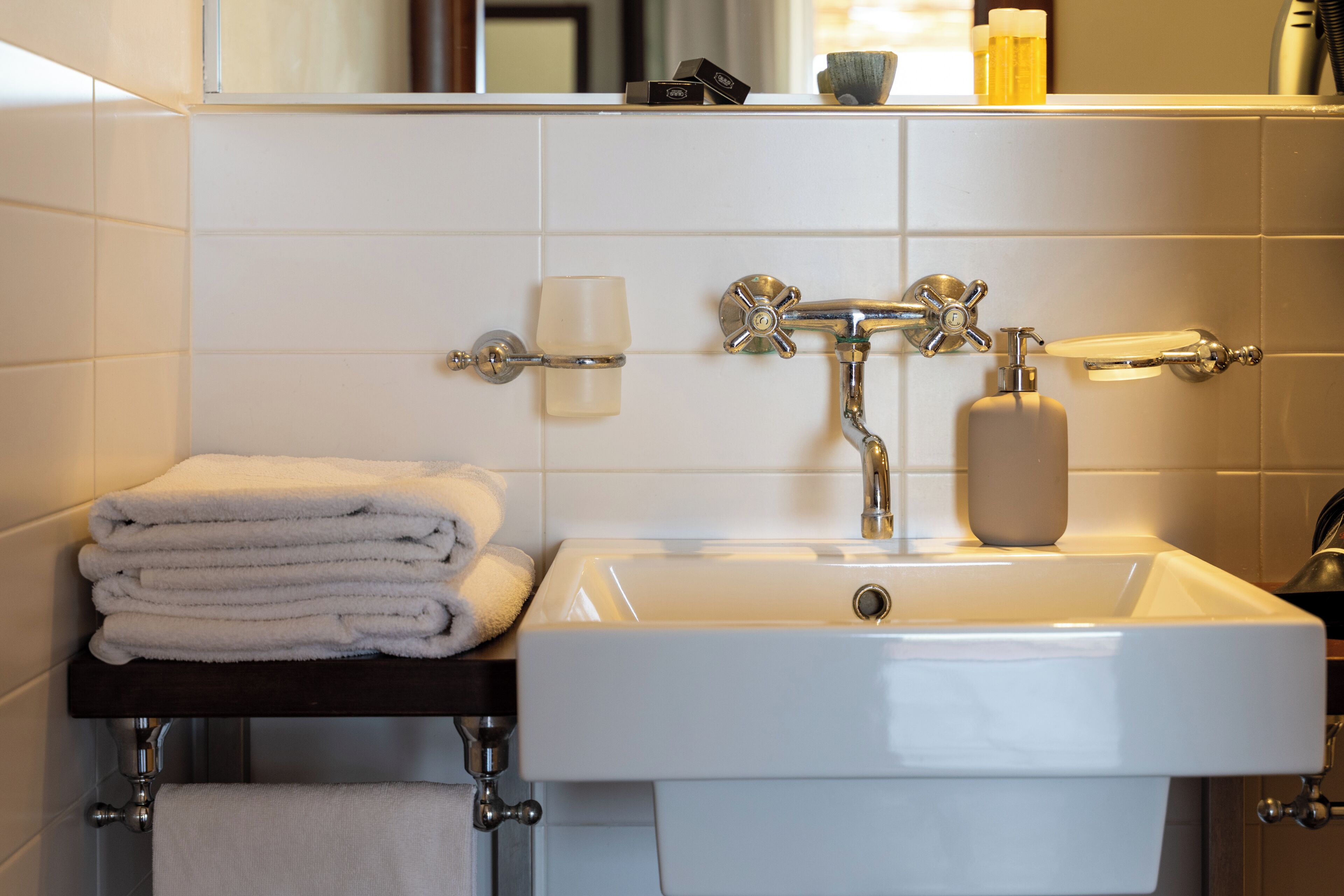 Un bagno con un lavandino bianco, un portasciugamani con asciugamani piegati, un erogatore di sapone e uno specchio sopra il lavandino.