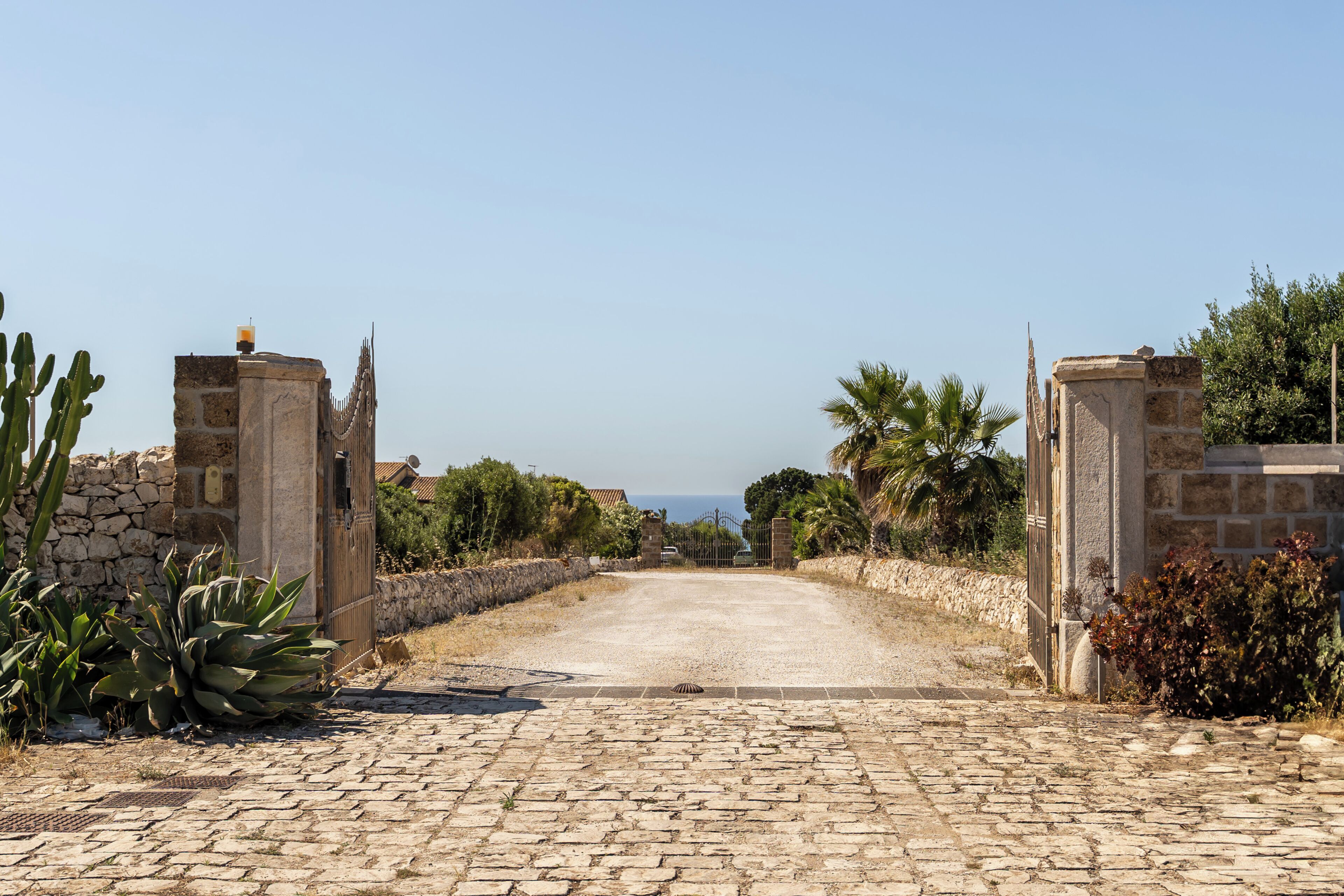 Un vialetto acciottolato conduce a un cancello in pietra, con vista sul mare in lontananza.