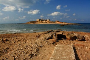Paesaggio costiero con spiaggia rocciosa, un sentiero in pietra e un edificio con un faro su una piccola isola sullo sfondo.
