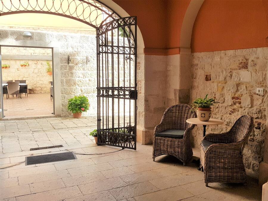 A stone-walled courtyard with wicker chairs and a small table, a metal gate, and a potted plant.
