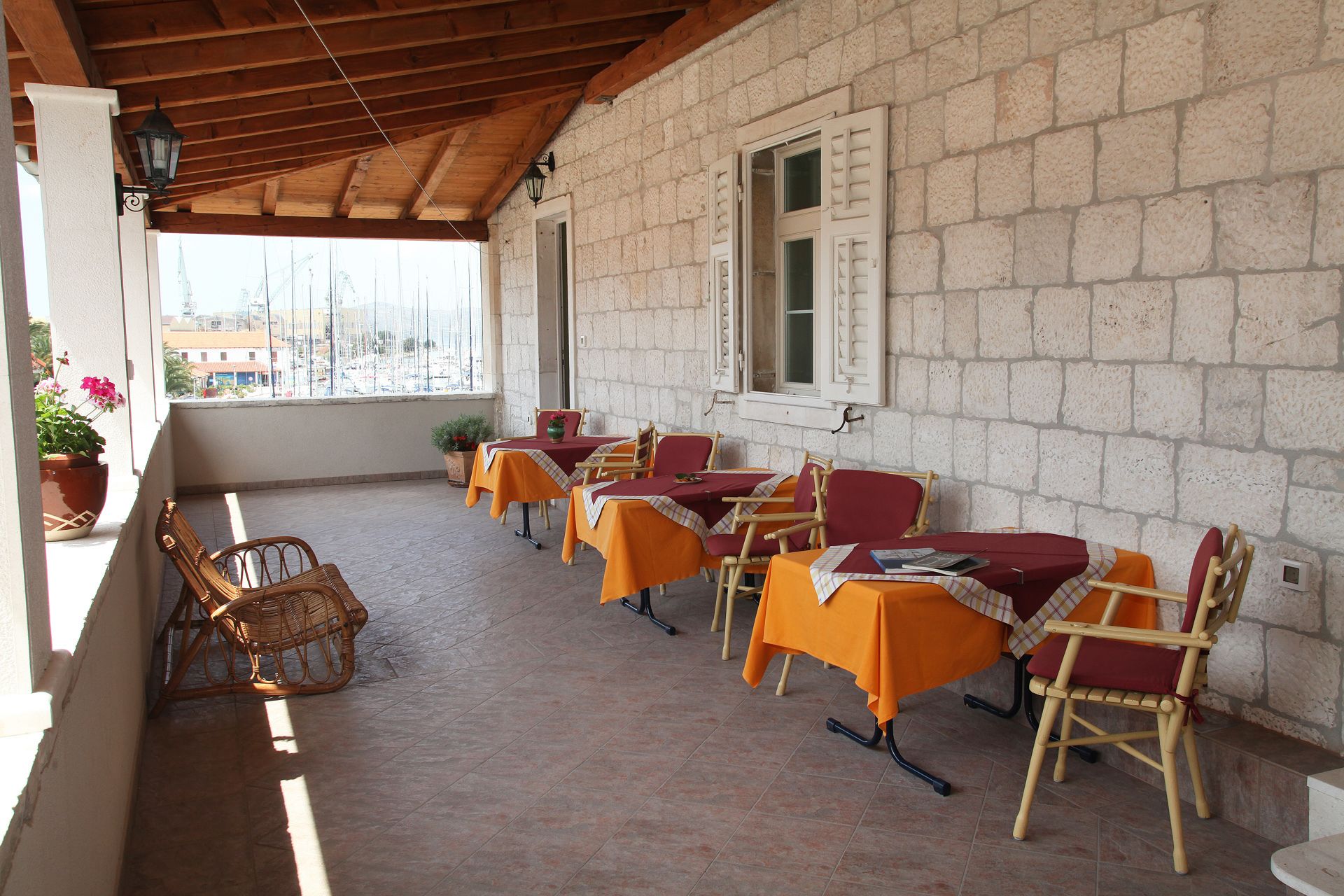 An outdoor patio with stone walls, wooden beams, and tables with orange tablecloths and wooden chairs.