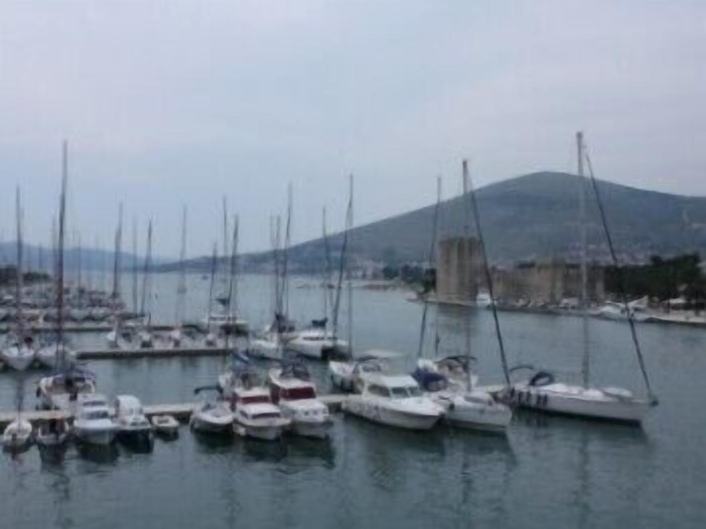 A marina with multiple boats docked, a mountainous backdrop, and a clear sky.
