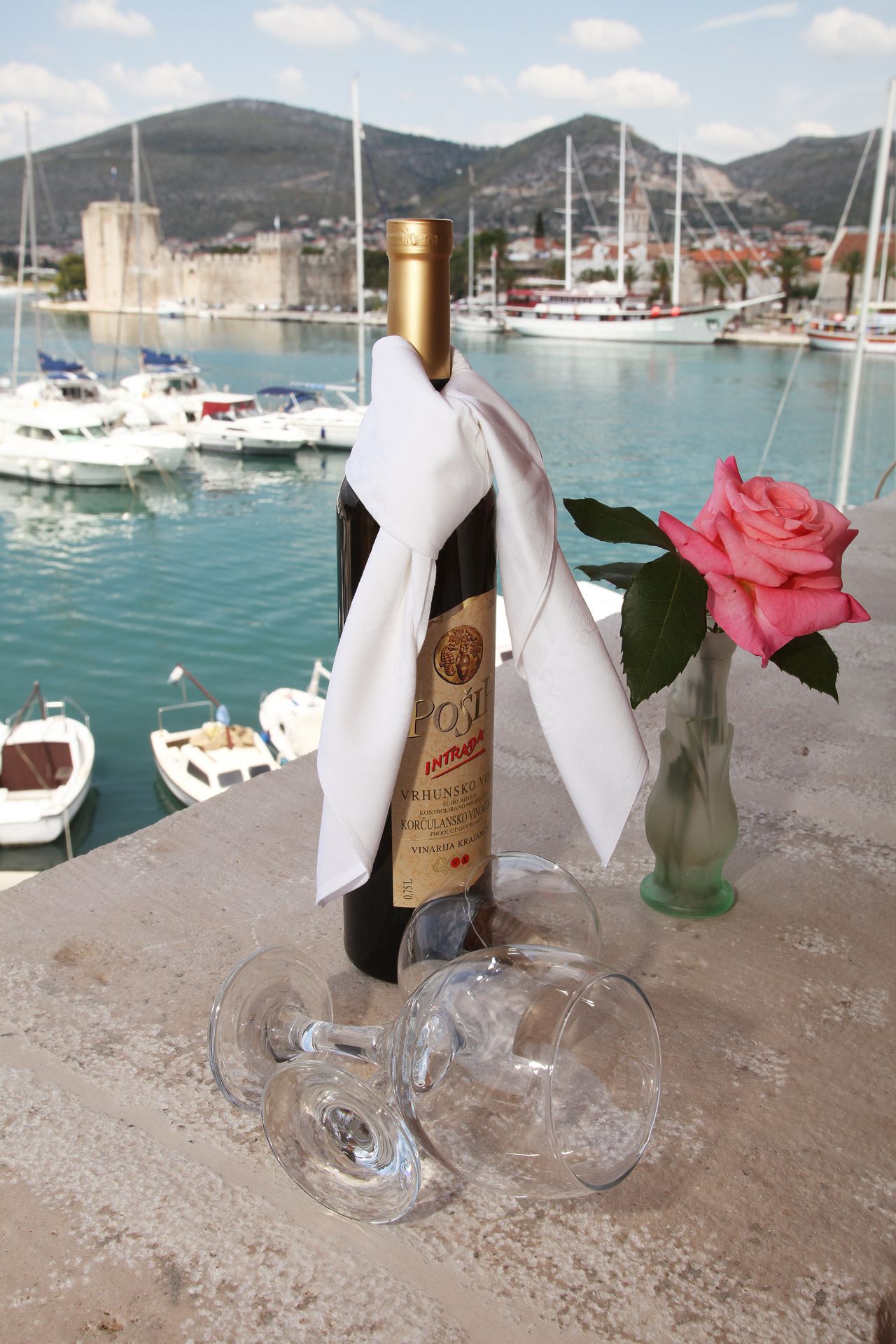 A bottle of wine with a white cloth tied around it, placed on a stone surface with two empty wine glasses beside it. A pink rose is in a clear vase to the right. In the background, there is a marina with boats and a mountainous landscape.