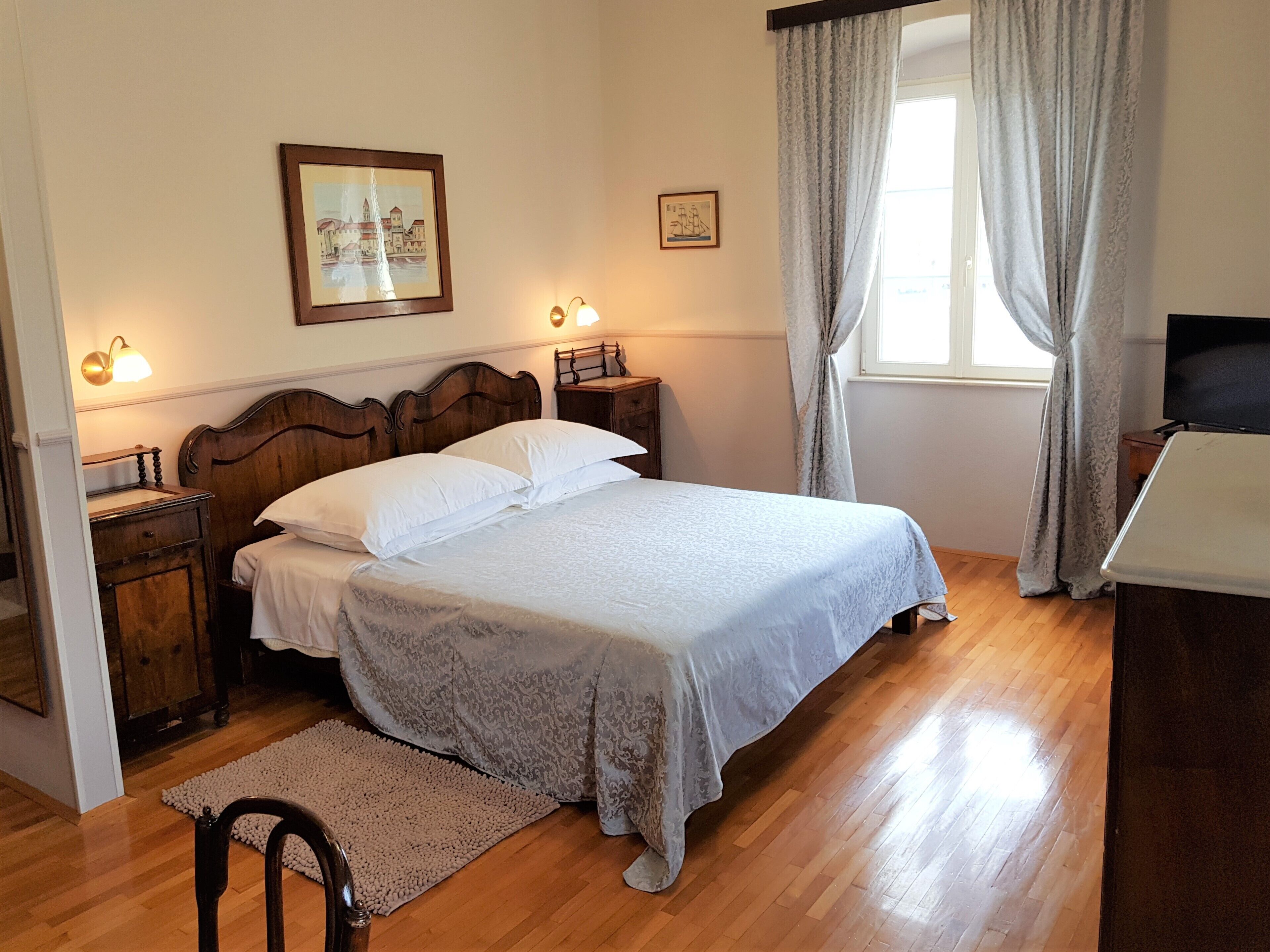 A neatly arranged bedroom with a wooden bed, two bedside tables, a nightstand, a framed picture on the wall, and a window with curtains.