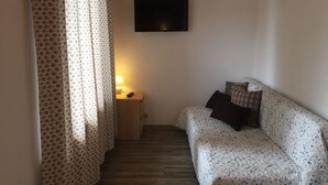 A room with a patterned sofa, a small bedside table with a lamp, and a TV mounted on the wall. - Pension Waldkrieber (Hermagor-Pressegger See)