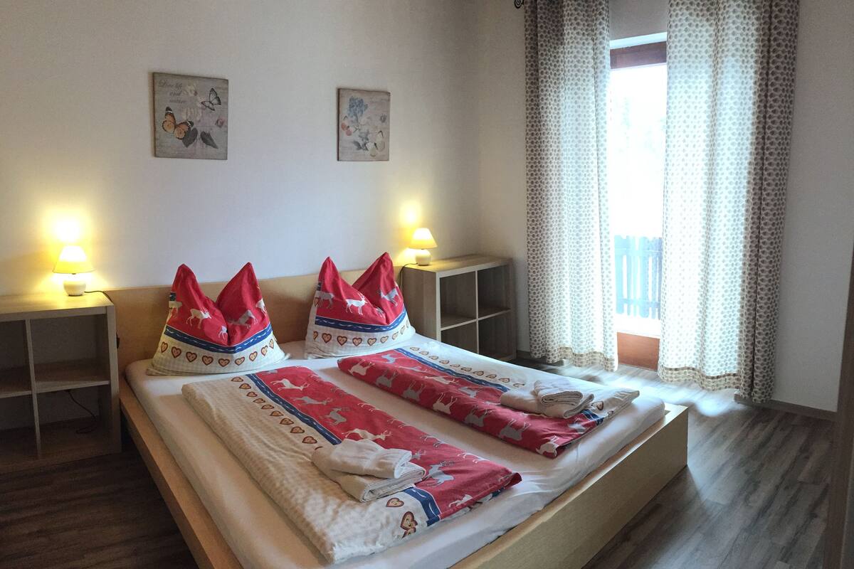 A hotel room with a wooden bed, two bedside tables with lamps, a window with patterned curtains, and two wall-mounted pictures.
