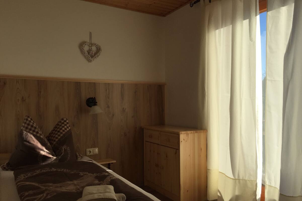 A bedroom with a wooden headboard, a bed with a patterned bedspread, a nightstand, a small wooden cabinet, and a window with sheer curtains.