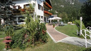 A building with a sign reading "Pension Waldkriiber". It has a paved entrance, outdoor seating with umbrellas, and is surrounded by greenery and mountains in the background. - Pension Waldkrieber (Hermagor-Pressegger See)