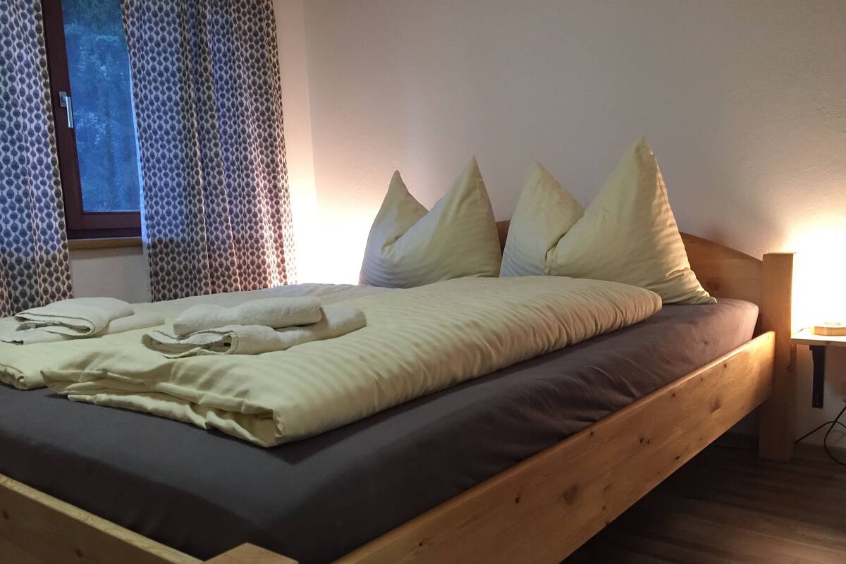 A wooden bed with a dark mattress, white pillows, and a neatly folded white towel.