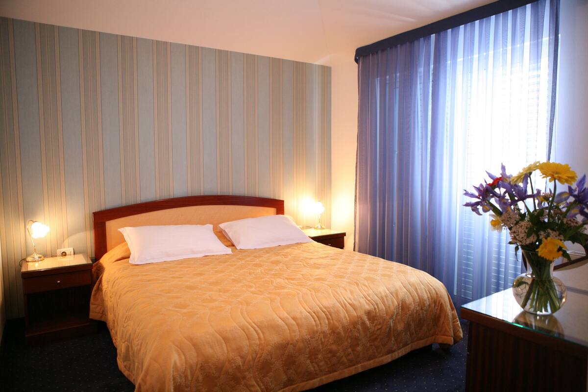 Hotellihuone, jossa on suuri sänky, kaksi yöpöytälamppua, yöpöytä, maljakko kukkineen ja ikkuna, jossa on verhot.