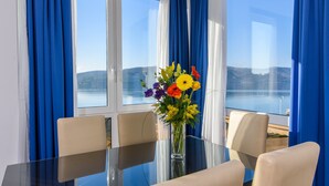 Un coin repas avec une table en verre, quatre chaises, un vase de fleurs et une vue sur la mer, grâce à de grandes fenêtres ornées de rideaux bleus.