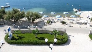 Un hôtel en bord de mer doté d’une plage aménagée avec des transats et des parasols, offrant une vue dégagée sur la mer et comprenant un jardin paysager avec des palmiers.