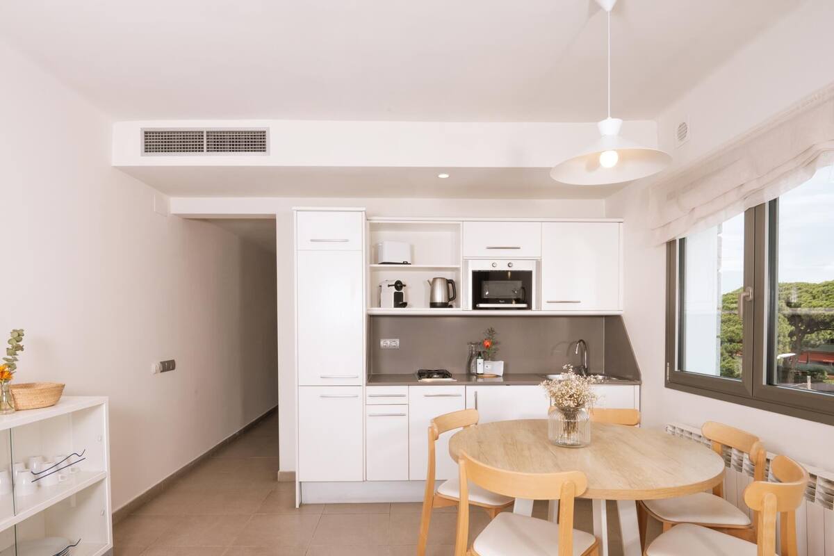 A modern kitchen with white cabinets, a microwave, and a sink. There is a dining table with four chairs and a vase of flowers.