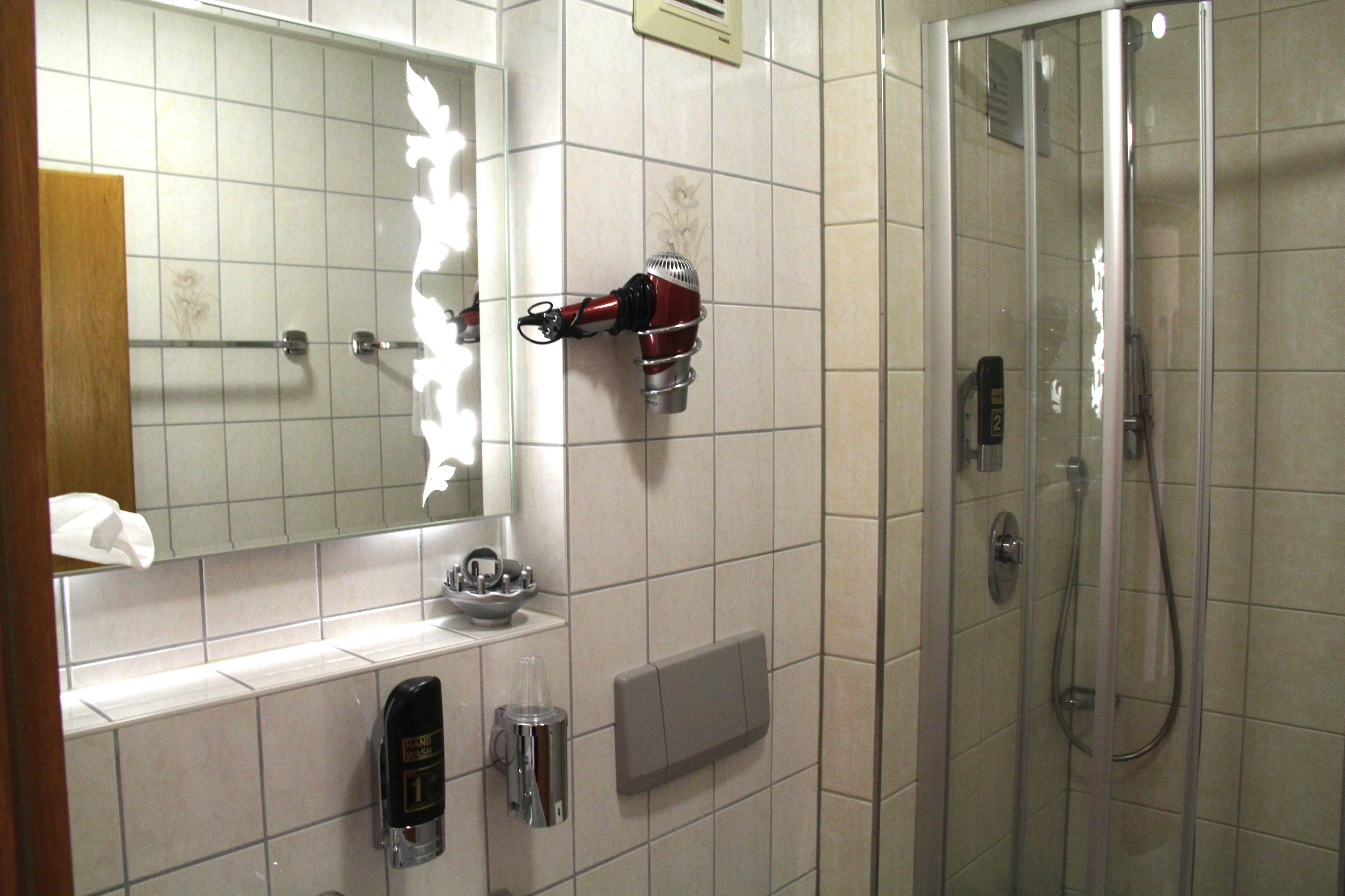 A bathroom with a mirror, a hairdryer mounted on the wall, a soap dispenser, and a shower area.