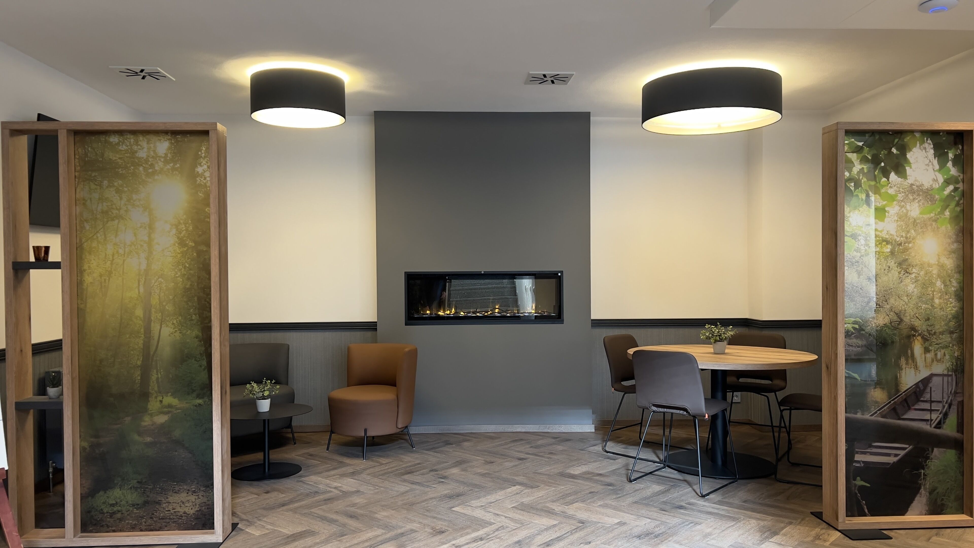 Een moderne kamer met een open haard, twee hanglampen, een ronde tafel met stoelen en een groot raam met uitzicht op bomen.