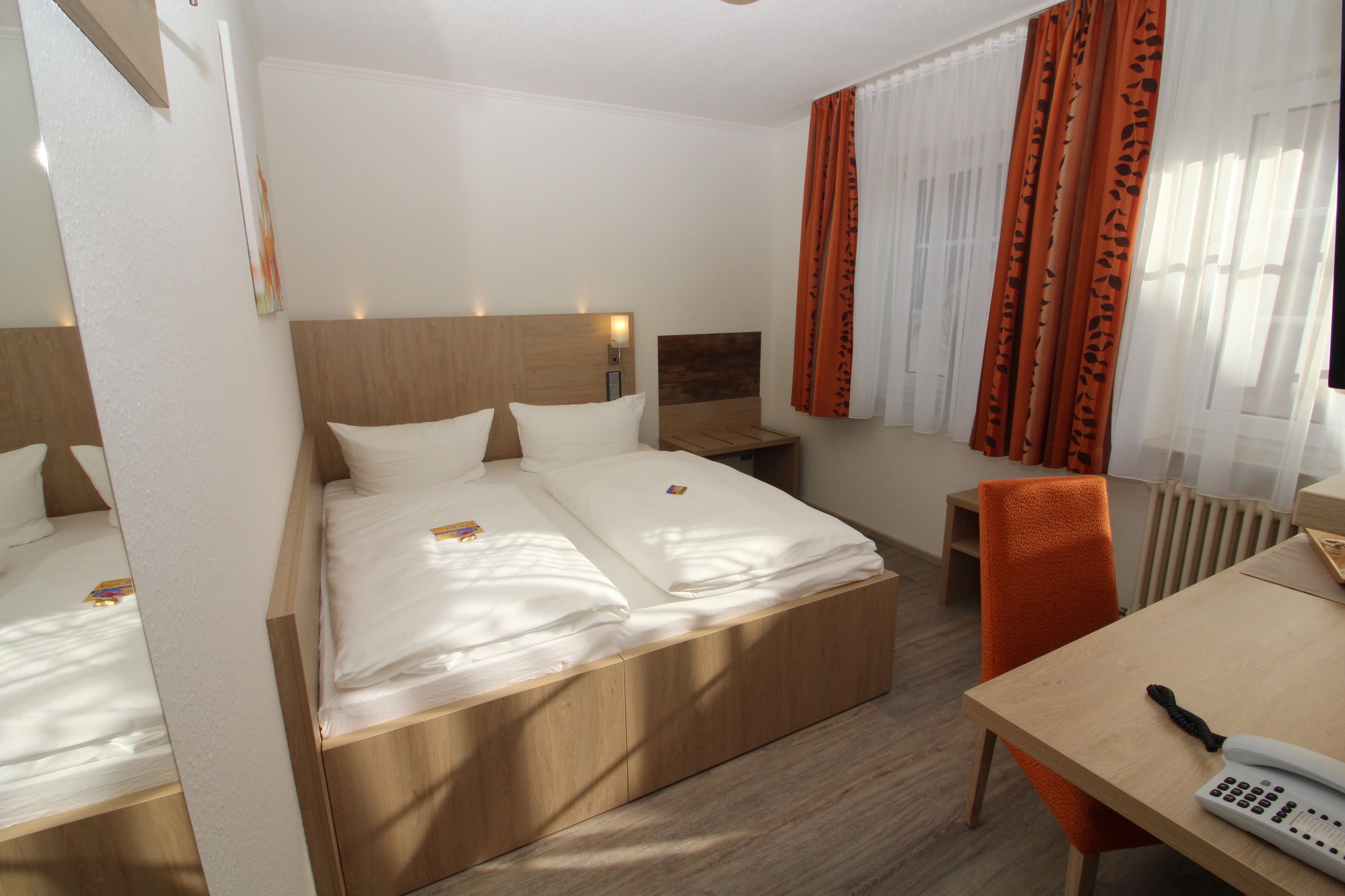 A hotel room with a double bed, a wooden headboard, a bedside table with a lamp, an orange chair, and a desk with a telephone.