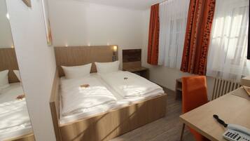 A hotel room with a double bed, a wooden headboard, a bedside table with a lamp, an orange chair, and a desk with a telephone.
