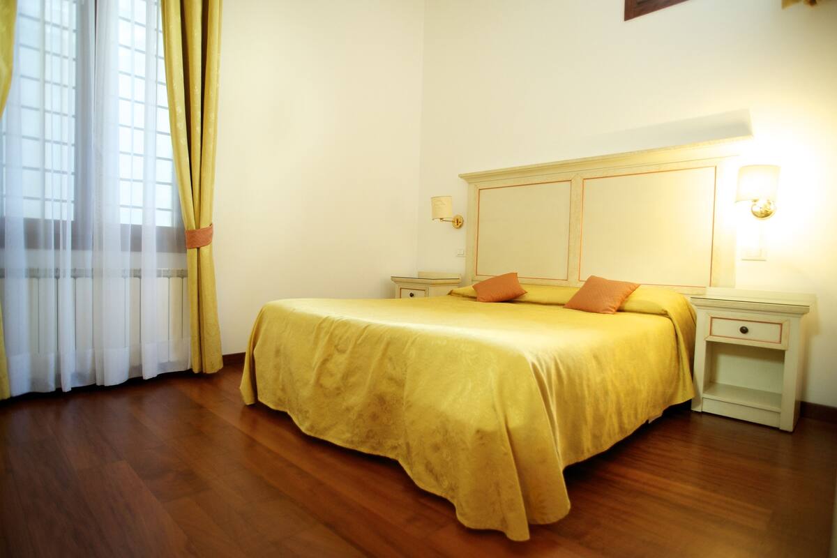 A room with a large bed, two bedside tables, a nightstand, and a window with curtains.