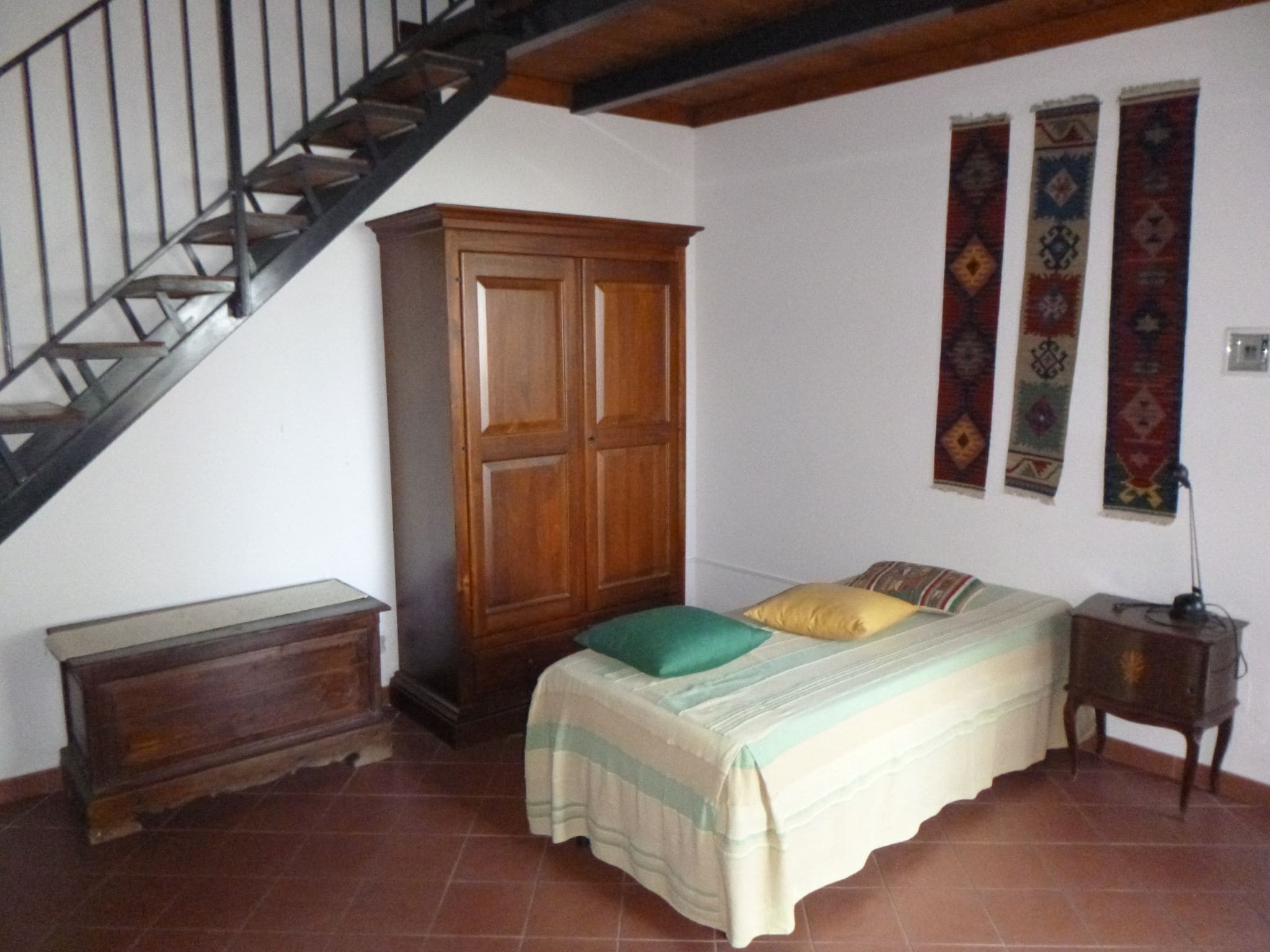 A bedroom with a bed, a wooden wardrobe, a nightstand, a lamp, a staircase, and decorative wall hangings.