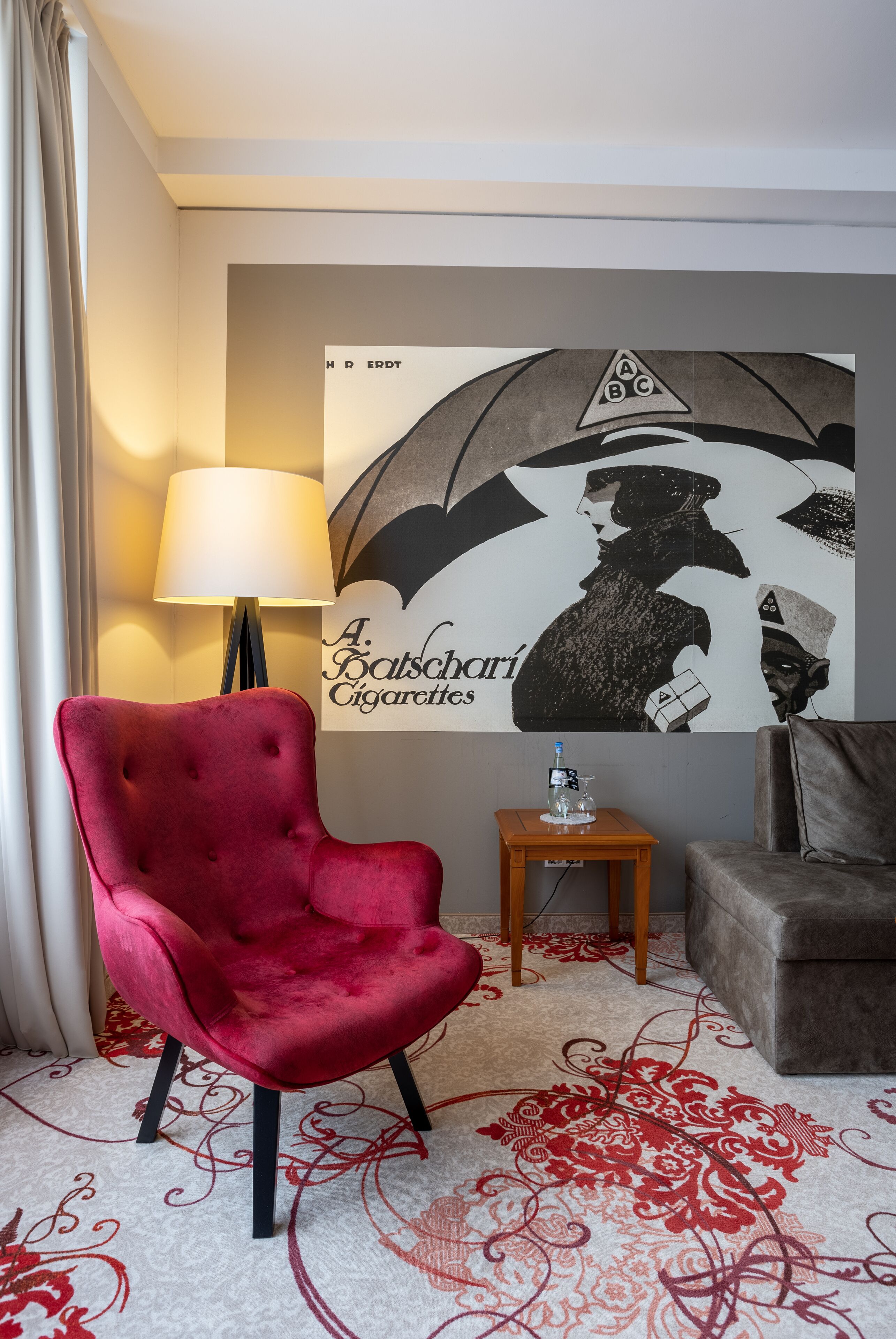 A room with a red velvet armchair, a grey sofa, a small wooden side table with a lamp, and a wall with a large poster.