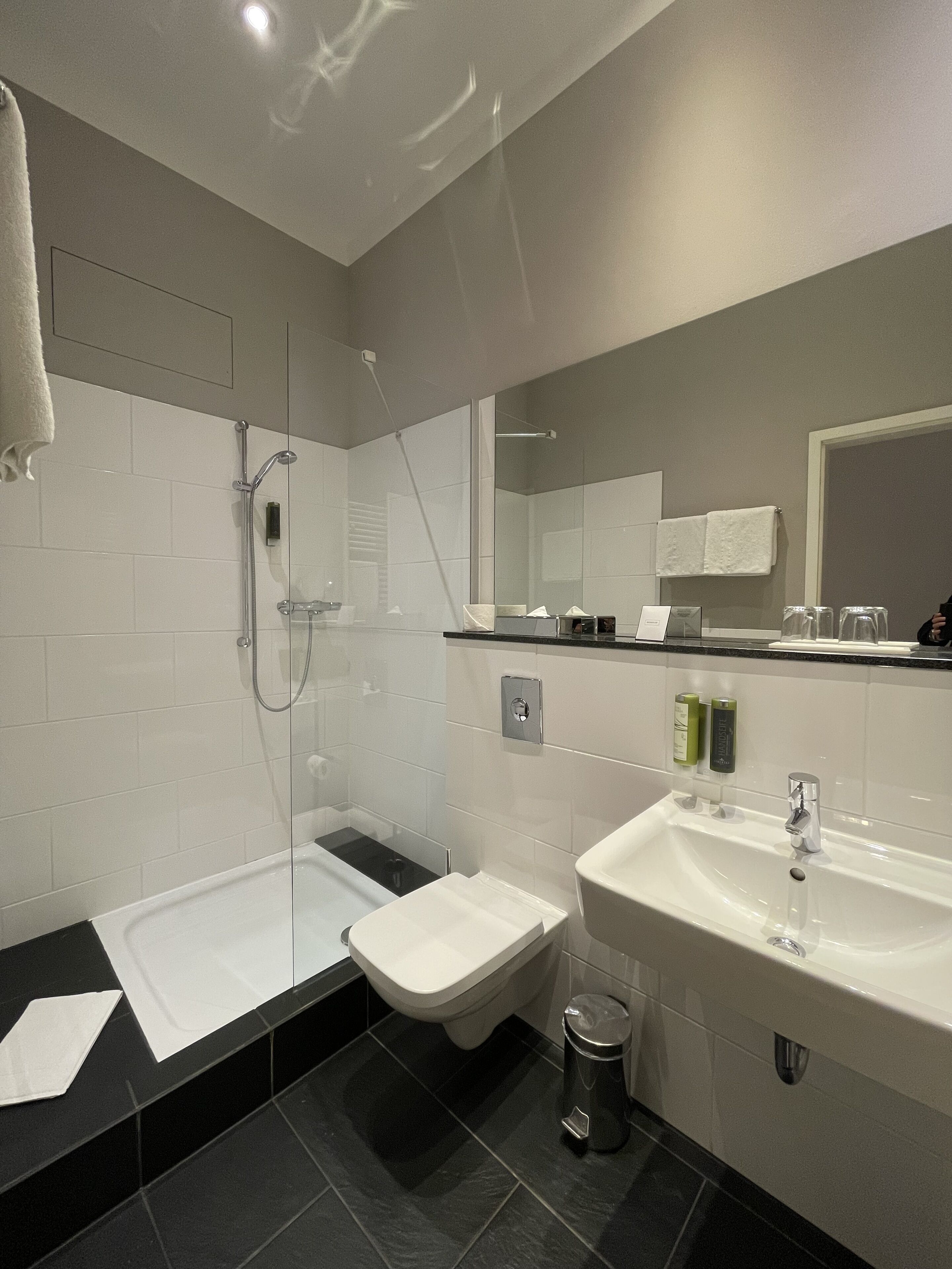 Une salle de bain moderne avec une douche à parois de verre, des toilettes blanches, un lavabo et une poubelle.