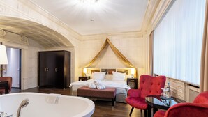 Habitación de hotel con una cama grande, una zona de estar con sillones rojos, una bañera y un ventanal amplio.