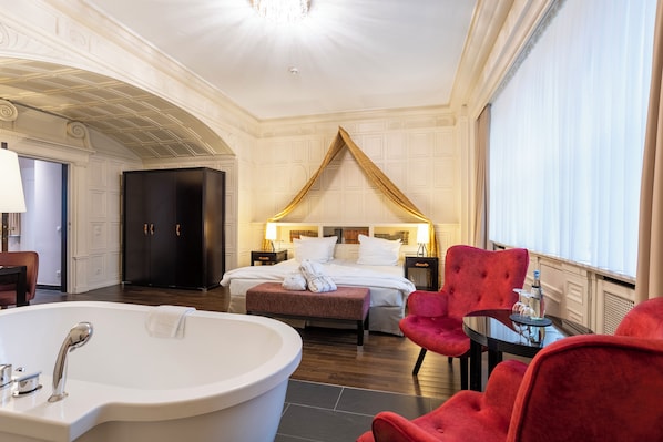 Une chambre d’hôtel avec un grand lit, un coin salon avec des fauteuils rouges, une baignoire et une grande fenêtre.