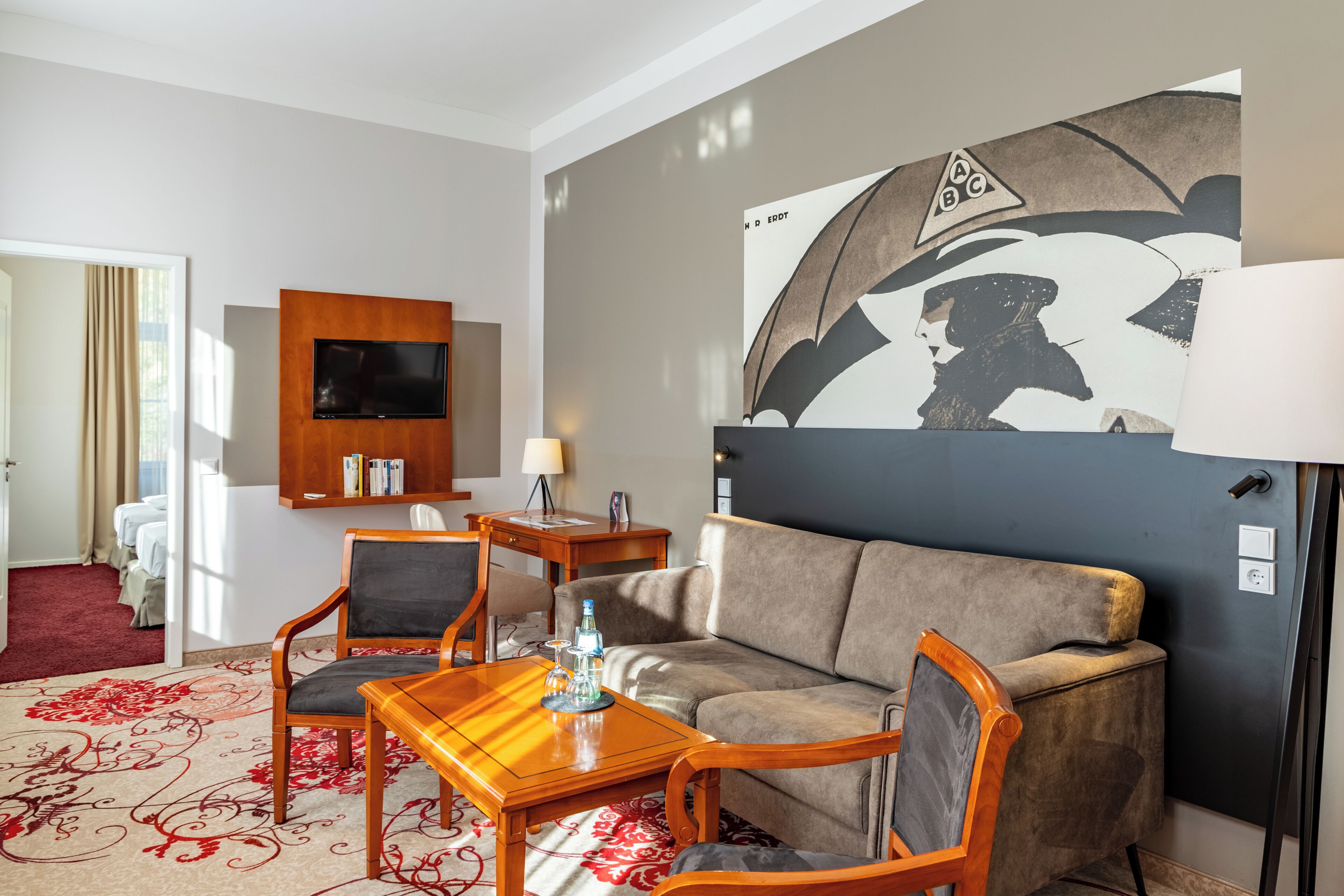A hotel room with a sofa, two armchairs, a wooden coffee table, a flat-screen TV, a wall-mounted shelf, and a red patterned carpet.
