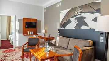 A hotel room with a sofa, two armchairs, a wooden coffee table, a flat-screen TV, a wall-mounted shelf, and a red patterned carpet.