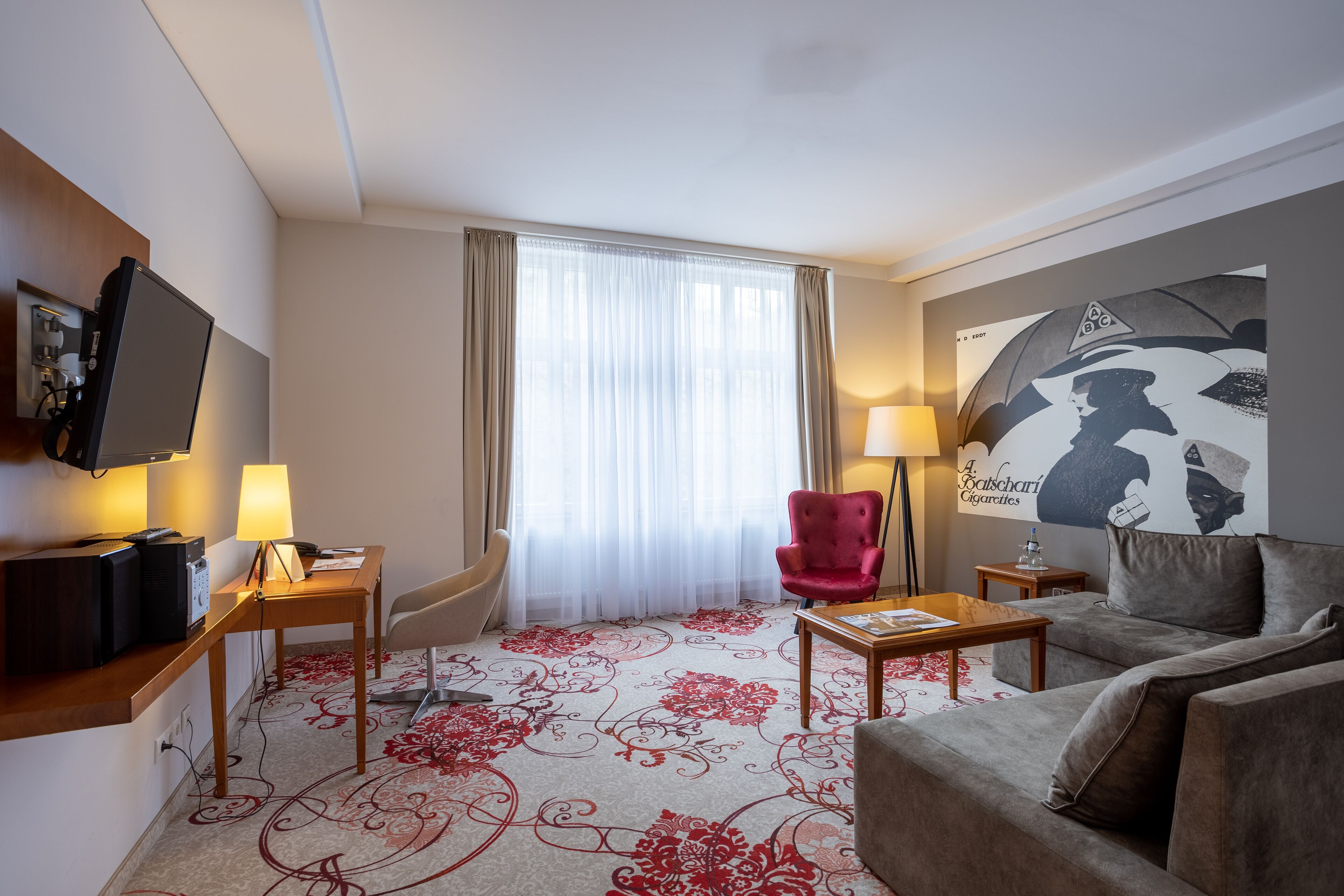 Habitación de hotel con televisor de pantalla plana, un escritorio con lámpara, un sillón rojo, un sofá y un ventanal con cortinas translúcidas.