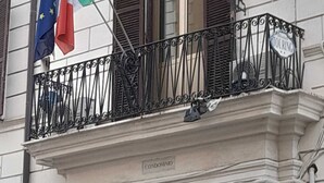 Un balcón de hotel con barandilla de hierro forjado, un cartel que dice 'Hotel Touring', y banderas de la Unión Europea e Italia.