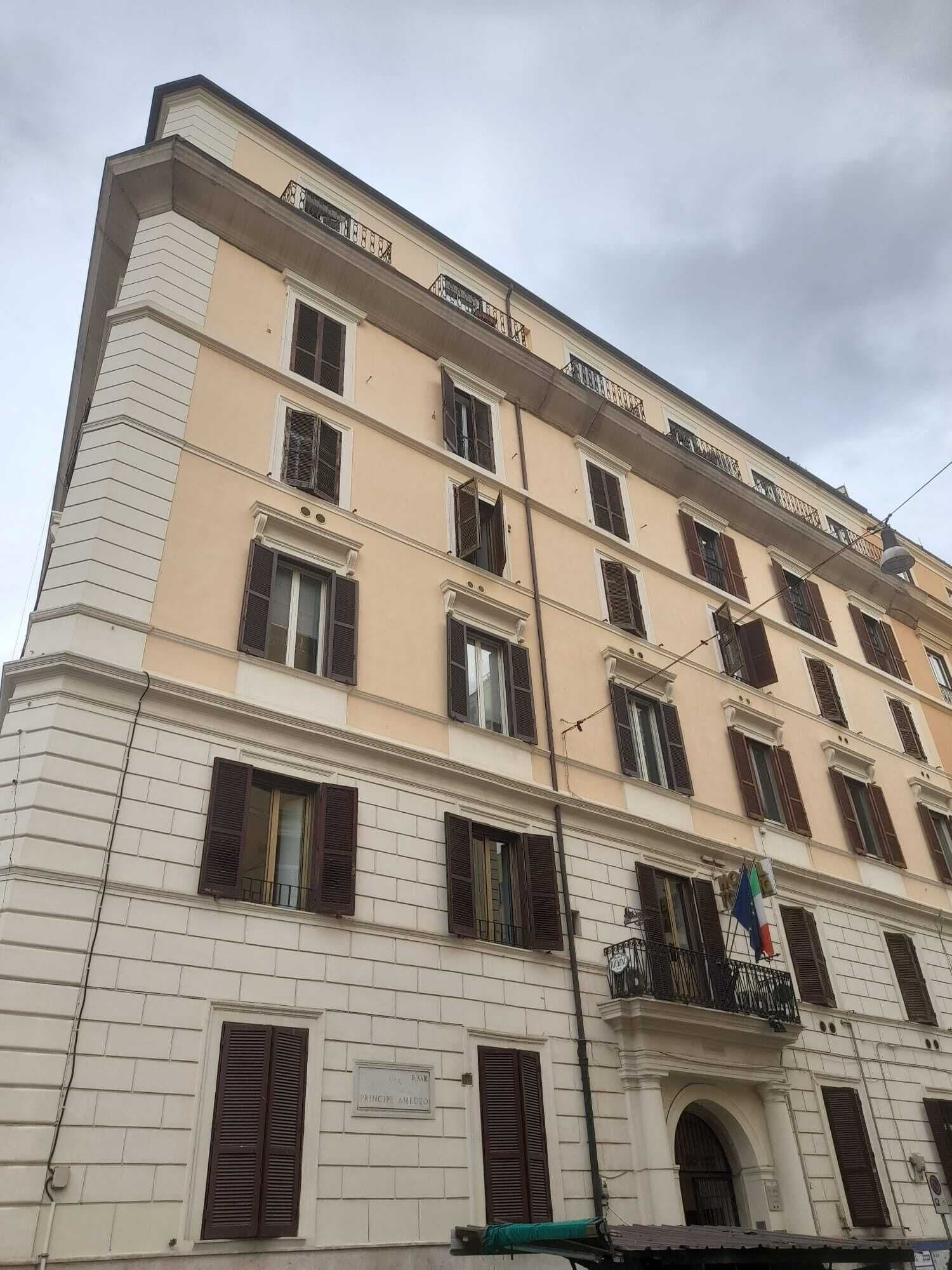 A multi-story building with a beige facade, multiple windows with shutters, and a balcony with a metal railing.