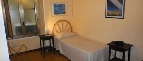 A small, single-bed room with a wooden floor, a bedside table, a nightstand, and a window with blinds.