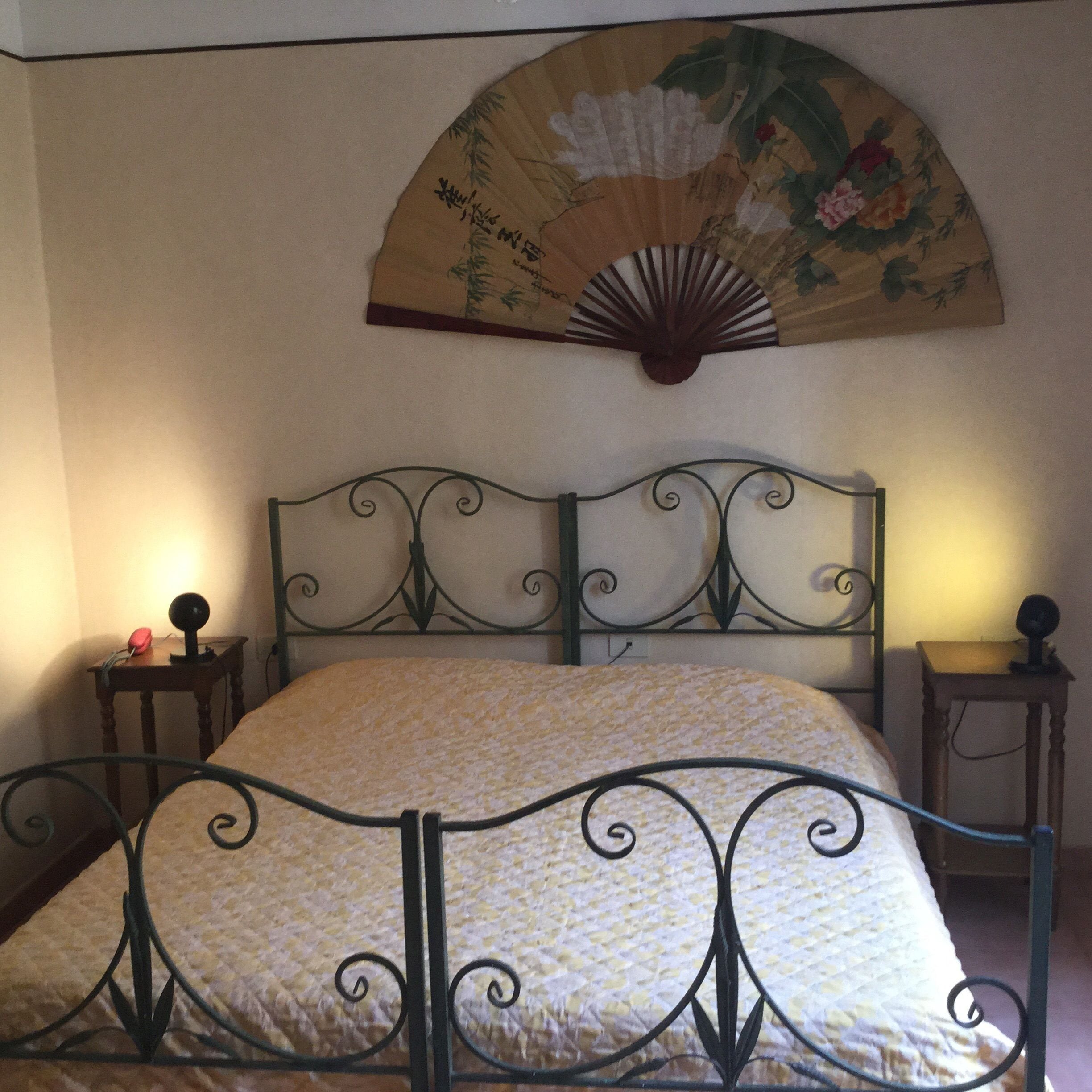 A bed with a metal frame and a patterned bedspread, a wall-mounted fan with floral design, and two bedside tables with lamps.