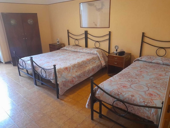 A bedroom with two metal-framed beds, a wooden wardrobe, a small bedside table, and a wall-mounted light fixture.