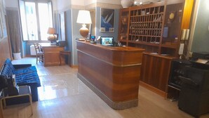 Un vestíbulo de hotel con un mostrador de recepción de madera, una zona de estar con sofás azules, una mesa de comedor y una cocina con estufa.