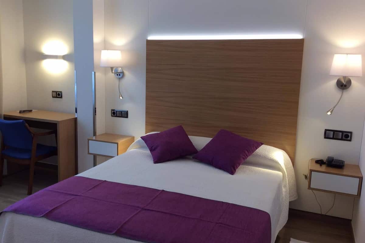 Quarto de hotel com uma cama, uma escrivaninha e duas mesas de cabeceira.